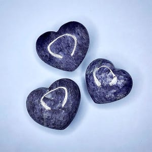 Puede incluir: Tres piedras en forma de coraz&oacute;n en tonos morados y azules est&aacute;n dispuestas sobre una superficie azul claro. Cada piedra tiene una marca blanca curvada. Las piedras tienen una textura pulida y suave, y la est&eacute;tica general es minimalista y elegante.