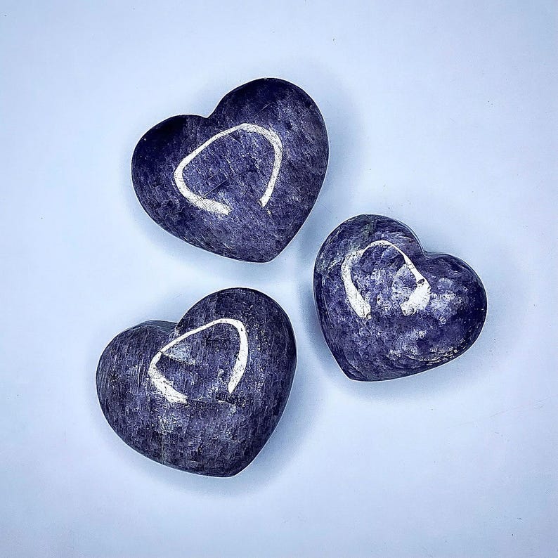 Puede incluir: Tres piedras en forma de coraz&oacute;n en tonos morados y azules est&aacute;n dispuestas sobre una superficie azul claro. Cada piedra tiene una marca blanca curvada. Las piedras tienen una textura pulida y suave, y la est&eacute;tica general es minimalista y elegante.
