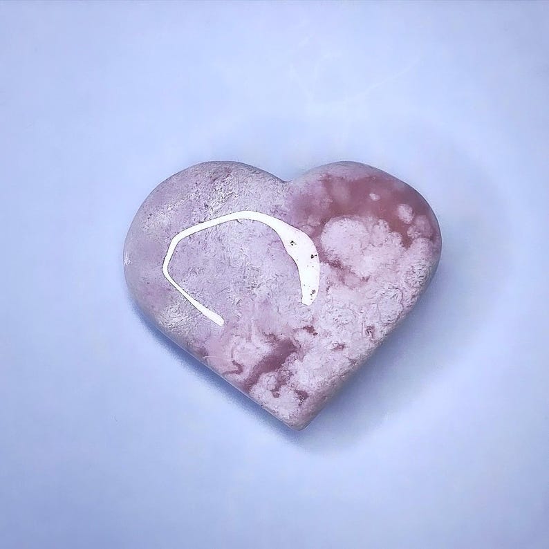 May include: A heart-shaped stone with a pink and white marbled pattern. A white, curved design is on the top of the heart. The stone is set against a light blue background.