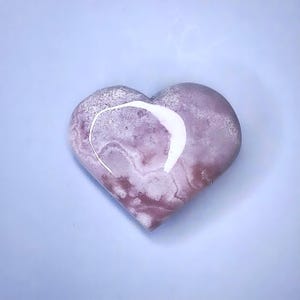 May include: A heart-shaped stone with a marbled appearance in shades of pink and white. A white, curved line is on the top of the heart. The stone is smooth and polished, and it sits on a light blue surface.