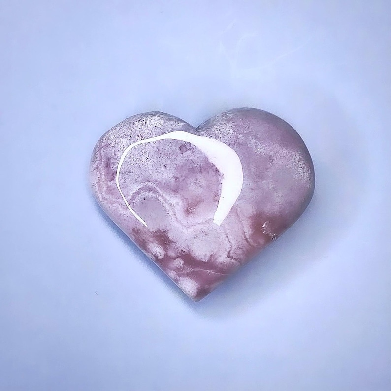 May include: A heart-shaped stone with a marbled appearance in shades of pink and white. A white, curved line is on the top of the heart. The stone is smooth and polished, and it sits on a light blue surface.