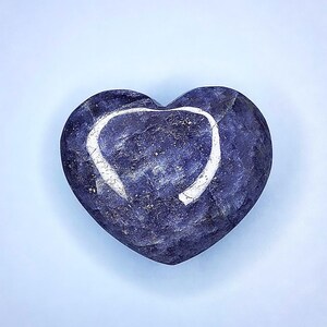 Puede incluir: Una piedra pulida en forma de coraz&oacute;n, en tonos azul profundo y morado. Un contorno blanco en forma de coraz&oacute;n es visible en la superficie. La piedra tiene una textura veteada sobre un fondo azul claro.