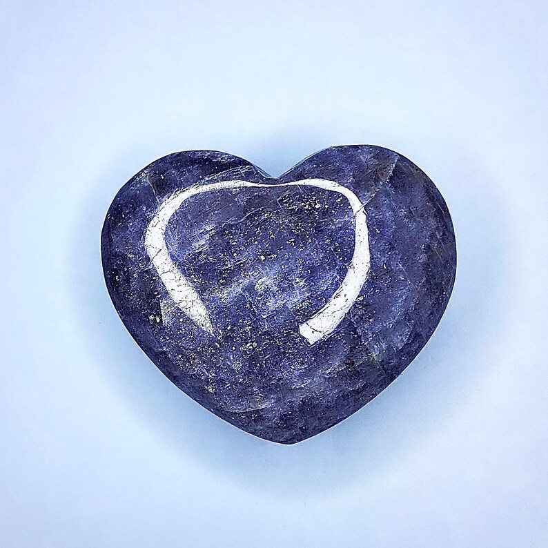 Puede incluir: Una piedra pulida en forma de coraz&oacute;n, en tonos azul profundo y morado. Un contorno blanco en forma de coraz&oacute;n es visible en la superficie. La piedra tiene una textura veteada sobre un fondo azul claro.