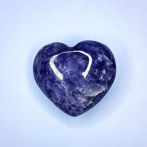 Puede incluir: Una piedra en forma de coraz&oacute;n, azul oscuro, con un dise&ntilde;o blanco curvado. La piedra tiene una superficie pulida y un aspecto natural texturizado. El coraz&oacute;n est&aacute; centrado en un fondo azul claro.