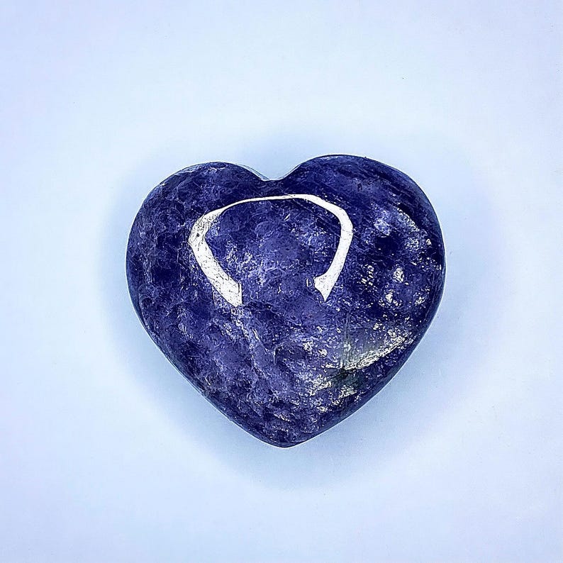 Puede incluir: Una piedra en forma de coraz&oacute;n, azul oscuro, con un dise&ntilde;o blanco curvado. La piedra tiene una superficie pulida y un aspecto natural texturizado. El coraz&oacute;n est&aacute; centrado en un fondo azul claro.
