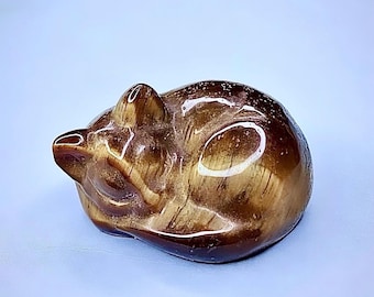 May include: A polished tiger's eye gemstone carved into the shape of a curled-up sleeping cat. The stone features a smooth, glossy finish with characteristic golden-brown chatoyant bands and natural mineral striations throughout the feline form.