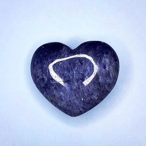 Puede incluir: Una piedra en forma de coraz&oacute;n, azul oscuro, con un dise&ntilde;o de c&iacute;rculo blanco incompleto. La piedra tiene una superficie texturizada y est&aacute; sobre un fondo azul claro. El objeto es un art&iacute;culo decorativo.