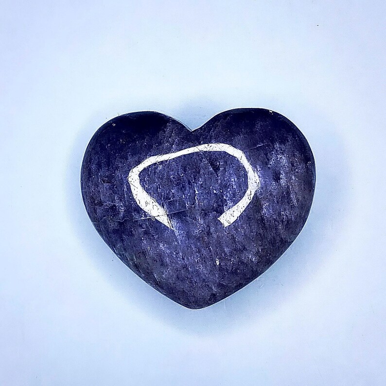 Puede incluir: Una piedra en forma de coraz&oacute;n, azul oscuro, con un dise&ntilde;o de c&iacute;rculo blanco incompleto. La piedra tiene una superficie texturizada y est&aacute; sobre un fondo azul claro. El objeto es un art&iacute;culo decorativo.