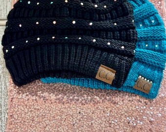 May include: Two knit beanies with rhinestones. One beanie is black and the other is teal. Both beanies have a brown leather tag with the letters "CC" on them.