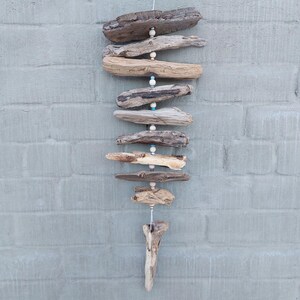 May include: A driftwood mobile with natural wood pieces and white and blue beads hanging from a string. The mobile is hanging against a grey brick wall.
