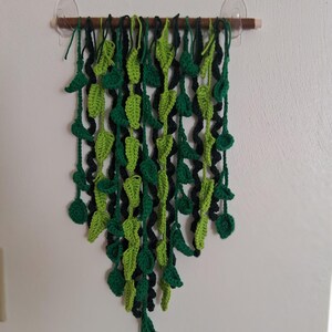 May include: A handmade crochet wall hanging featuring a cascade of green leaves. The leaves are in varying shades of green and black, attached to a wooden dowel. The wall hanging is secured with clear suction cups, creating a natural, organic aesthetic.
