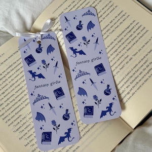 May include: Two light purple bookmarks with a fantasy theme, featuring illustrations of a dragon, book, rose, potion bottle, dagger, crown, and wings. The text "fantasy girlie" is printed on each bookmark. A white ribbon is attached.