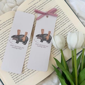 Peut inclure: Deux marque-pages illustrés représentant un hippopotame de dessin animé en train de lire un livre. Les marque-pages sont blancs avec des rubans roses et le texte "Ne m'interrompez pas pendant ma lecture !"