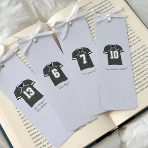 May include: Four light gray bookmarks with illustrations of football jerseys. Each jersey has a different number and a white ribbon tied at the top. Text on the bookmarks includes "nice legs", "I'm yours", and "no matter what".