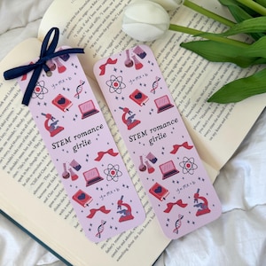 May include: Two pink bookmarks with a STEM romance theme. The bookmarks feature a variety of science-related illustrations, including a microscope, a DNA strand, a beaker, and a laptop. The text on the bookmarks reads "STEM romance girlie" and "y=mx+b".