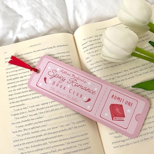 May include: Pink and white bookmark with a pink ticket design. The bookmark features a book with a red chili pepper on top and the text "Lifetime Membership Spicy Romance Book Club The steamier the better". The bookmark also features the text "Admit One" in a white circle with a red border.