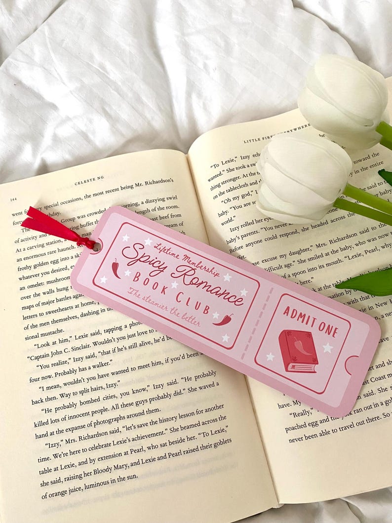 May include: Pink and white bookmark with a pink ticket design. The bookmark features a book with a red chili pepper on top and the text "Lifetime Membership Spicy Romance Book Club The steamier the better". The bookmark also features the text "Admit One" in a white circle with a red border.