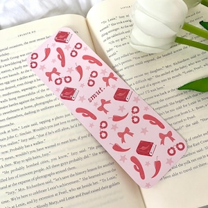 May include: A pink bookmark with a red book, handcuffs, chili peppers, and bows. The bookmark has the word "smut" printed on it.