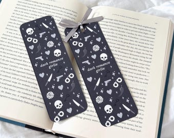 May include: Two dark grey bookmarks with a repeating pattern of skulls, guns, hearts, knives, handcuffs, chilli peppers, and roses. The text "dark romance girlie" is printed on each bookmark. A grey ribbon is attached to one.