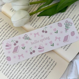 Könnte beinhalten: Ein rosa und weißes Lesezeichen mit einem floralen Design und dem Text "hello spring". Das Lesezeichen zeigt Kirschen, Schleifen, einen Korb und ein Buch.