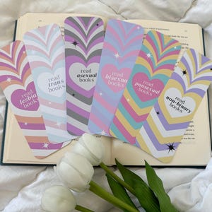 Puede incluir: Conjunto de seis marcapáginas coloridos con rayas arcoíris y corazones. Cada marcapáginas presenta el texto "leer libros [identidad]" con diferentes identidades representadas: lesbiana, trans, asexual, bisexual, pansexual y no binario.