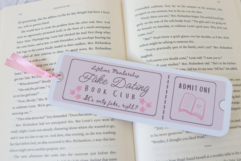 May include: A pink and white bookmark with the text "Lifetime Membership Fake Dating Book Club It's only fake, right?" and a pink ticket with the text "Admit One" and a pink book icon.