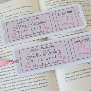 May include: Two pink and white bookmarks with the text "Lifetime Membership Fake Dating Book Club It's only fake, right?" and "ADMIT ONE" with a pink book icon. The bookmarks are on top of a book.