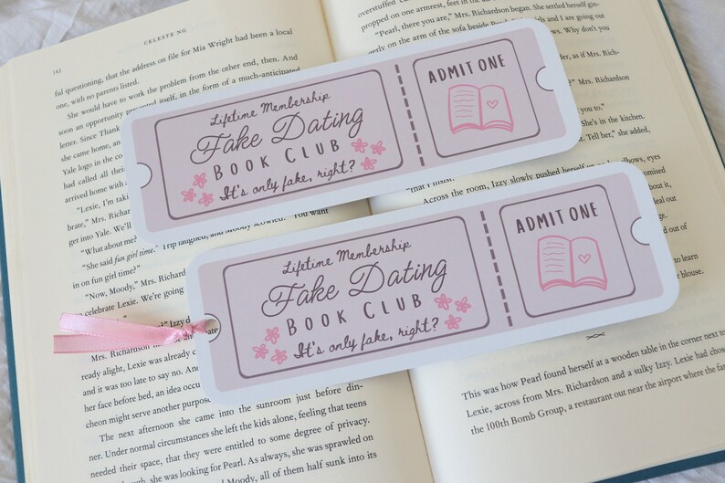 May include: Two pink and white bookmarks with the text "Lifetime Membership Fake Dating Book Club It's only fake, right?" and "ADMIT ONE" with a pink book icon. The bookmarks are on top of a book.