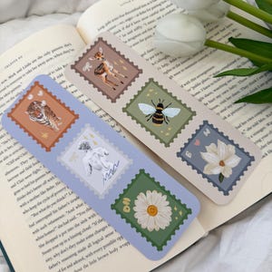 May include: A set of six bookmarks featuring different illustrations. Each bookmark is shaped like a postage stamp with a blue border and a white background. The illustrations include a deer, a bee, a woman, a daisy, a magnolia flower, and a tiger.