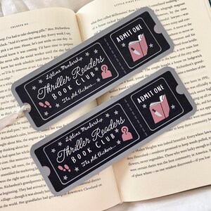 Op de afbeelding: Twee grijze en zwarte boekenleggertickets met een roze en witte illustratie van een boek en een mes. De tekst op de boekenleggers luidt "Lifetime Membership Thriller Readers Book Club... Het plot verdikt..." en "Toegang".