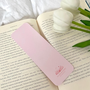 May include: A pink bookmark with a pink open book design and the text "Starcrossed Bookshop" in white. The bookmark is lying on a book with a white background.