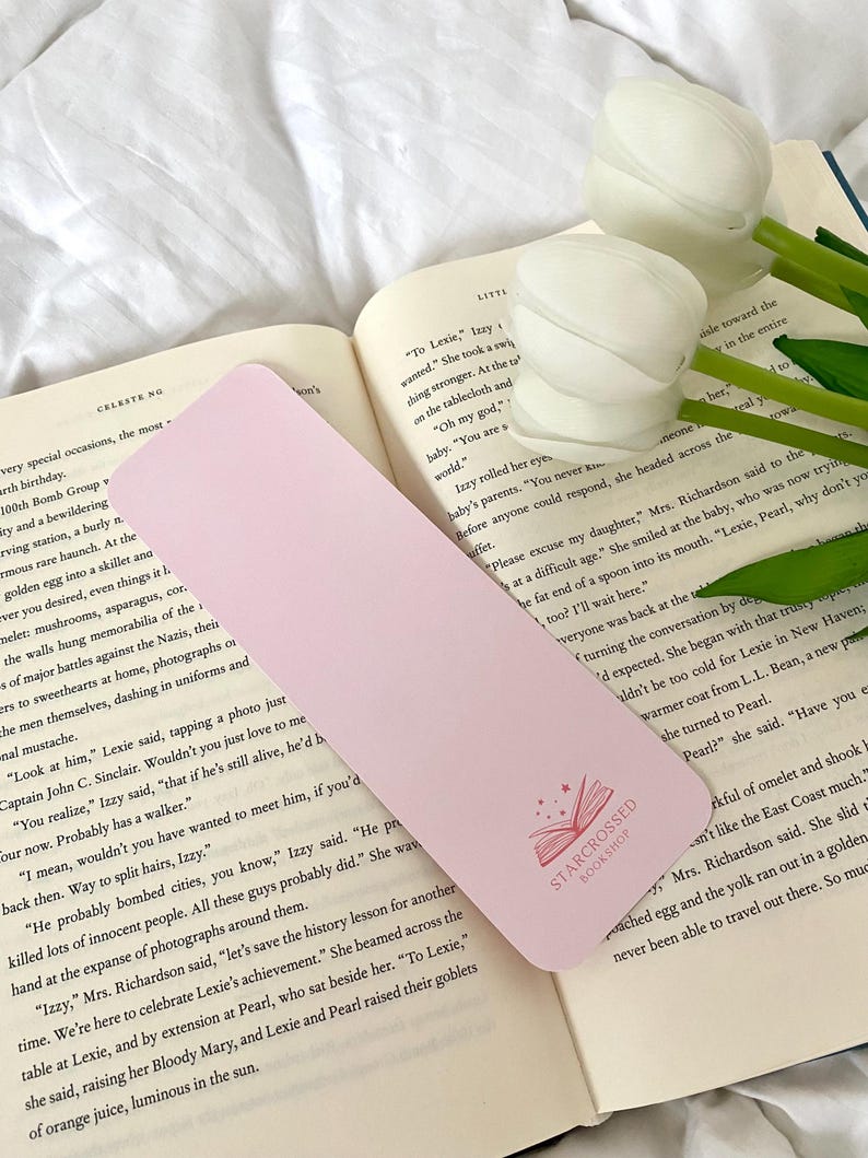 May include: A pink bookmark with a pink open book design and the text "Starcrossed Bookshop" in white. The bookmark is lying on a book with a white background.