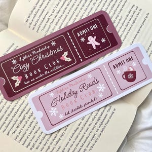 May include: Two pink and burgundy bookmark tickets with a winter holiday theme. The top bookmark says "Lifetime Membership Cozy Christmas Book Club kiss me under the mistletoe..." and the bottom bookmark says "Lifetime Membership Holiday Reads Book Club hot chocolate provided!" Both bookmarks have a ticket design with the words "Admit One" printed on them.