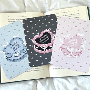 May include: Three printable bookmarks with a heart-shaped design and a pattern of small hearts. The bookmarks feature the text "Probably reading fantasy", "Probably reading dark romance", and "Probably reading romance".