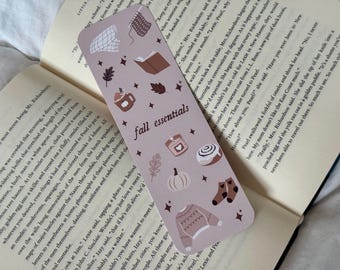 Puede incluir: Un marcapáginas rectangular con un fondo malva claro, con ilustraciones temáticas de otoño. El diseño incluye una taza de café, un libro abierto, una calabaza, un suéter, calcetines y el texto "fall essentials".