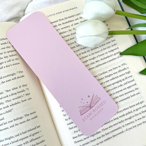 May include: A light purple bookmark featuring a white star and a book design. The bookmark has the text "Starcrossed Bookshop" printed on it.