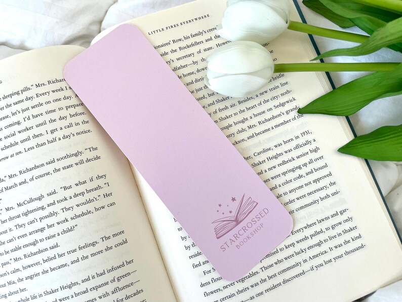 May include: A light purple bookmark featuring a white star and a book design. The bookmark has the text "Starcrossed Bookshop" printed on it.
