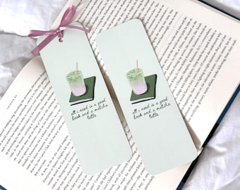 May include: Two light green bookmarks with a pink ribbon, each featuring a matcha latte illustration on a green book. The text "all I need is a good book and a matcha latte" is printed on each bookmark. The bookmarks are placed on an open book.