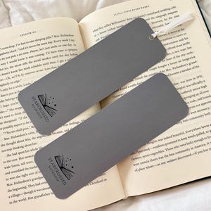 Op de afbeelding: Twee grijze bladwijzers met een zwart-witte illustratie van een boek met sterren. De tekst "Starcrossed Bookshop" staat op elke bladwijzer gedrukt.