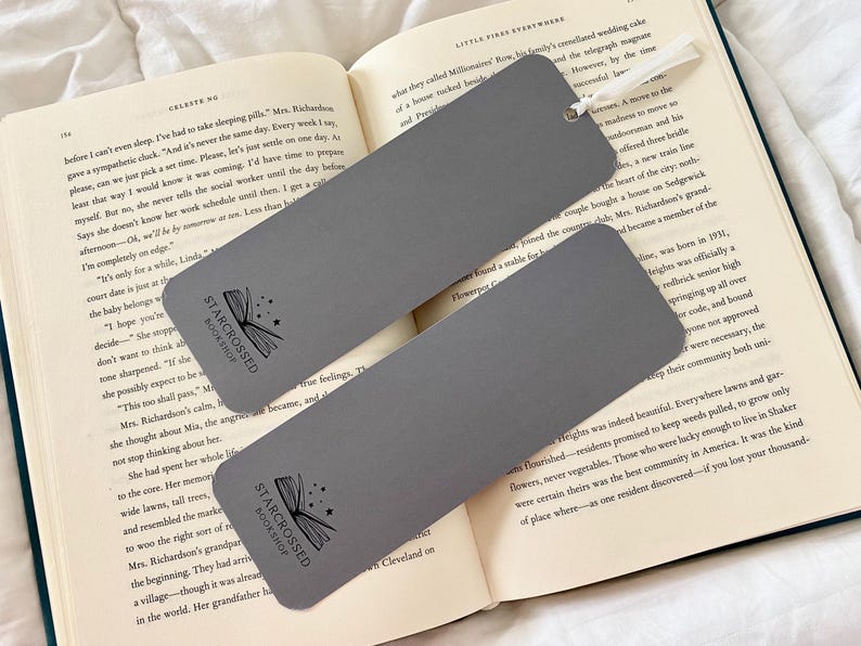 Op de afbeelding: Twee grijze bladwijzers met een zwart-witte illustratie van een boek met sterren. De tekst "Starcrossed Bookshop" staat op elke bladwijzer gedrukt.