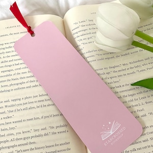 May include: A pink bookmark with a white outline of an open book and stars. The bookmark has the text "STARCROSSED BOOKSHOP" printed on it.