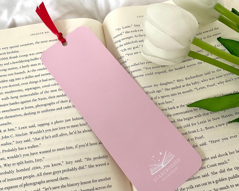 May include: A pink bookmark with a white outline of an open book and stars. The bookmark has the text "STARCROSSED BOOKSHOP" printed on it.