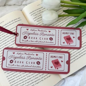 Pode incluir: Dois bilhetes de marcador de página rosa e branco com o texto "Membro vitalício Hopeless Romantic Book Club Apaixonamo-nos por personagens fictícios" e "Admitir um". Os marcadores de página apresentam um coração vermelho e um livro com um coração vermelho na capa.