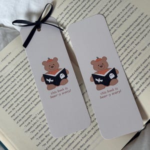 Può includere: Due segnalibri rettangolari color bianco sporco con un orso marrone che indossa un cappello a forma di zucca e legge un libro nero con un fantasma e un pipistrello bianchi. Il testo "this book is bear-y scary!" è stampato sotto l'orso. Un nastro nero.