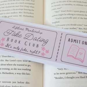 May include: A pink and white bookmark with the text "Lifetime Membership Fake Dating Book Club It's only fake, right?" and "ADMIT ONE" with a pink book illustration.