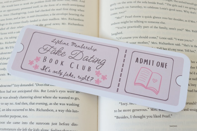 May include: A pink and white bookmark with the text "Lifetime Membership Fake Dating Book Club It's only fake, right?" and "ADMIT ONE" with a pink book illustration.