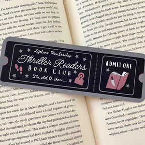 Op de afbeelding: Een zwarte en grijze bladwijzer met een ticketontwerp. De bladwijzer toont een boek met een mes dat eruit steekt en de tekst "Admit One". De andere kant van de bladwijzer heeft de tekst "Lifetime Membership Thriller Readers Book Club The plot thickens..." met een ster en een vraagteken.