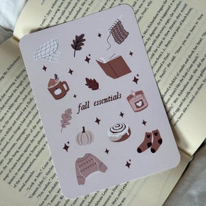Op de afbeelding: Een rechthoekige kaart met de tekst "fall essentials" en illustraties met herfstthema's, zoals een pompoen, een mok, een kaars en een trui. De kaart ligt op open boeken.
