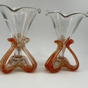 Puede incluir: Dos jarrones de cristal transparente con detalles en naranja. Los jarrones tienen una parte superior acampanada y con volantes y una base naranja con asas curvas y en bucle. El cristal tiene un aspecto ligeramente texturizado. Los jarrones son probablemente decorativos.