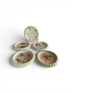 Könnte beinhalten: Eine Sammlung von fünf cremefarbenen Keramikplatten mit floralem Design. Die Teller haben einen gewellten Rand und zeigen ein zentrales Bouquet aus rosa, roten und gelben Blumen. Die Teller sind auf einer weißen Oberfläche angeordnet.